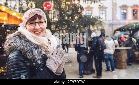Giovane donna che si gode un mercatino di Natale innevato, indossa abiti invernali e un berretto, sorridendo in macchina fotografica con un copyspace per il tuo testo individuale. Foto Stock