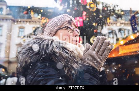 Donna con occhiali e un cappello invernale stringe le mani insieme per riscaldarsi, cadere la neve, mercatino di Natale Foto Stock