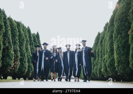 Gli studenti universitari multietnici in abiti da laurea celebrano con gioia il loro successo in un parco soleggiato. Foto Stock