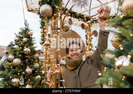 Happy boy che tiene in mano una confezione regalo e si diverte al mercatino vicino alle decorazioni natalizie Foto Stock