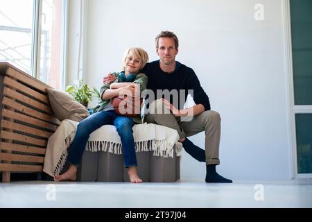 Figlio sorridente che tiene la pallacanestro e si siede con il padre a casa Foto Stock