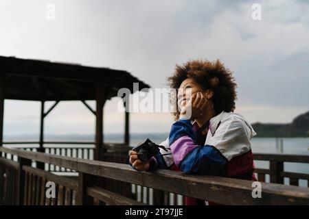 Donna felice e premurosa in piedi con la macchina fotografica vicino alla ringhiera Foto Stock