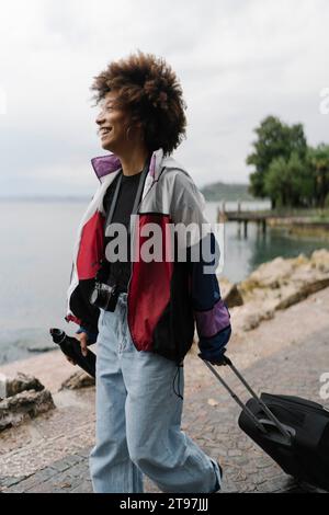 Giovane donna felice che cammina con la valigia vicino al lago Foto Stock