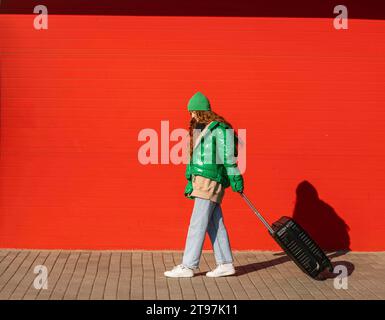 Giovane donna che indossa una giacca che cammina con una valigia davanti al muro rosso Foto Stock