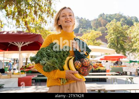 Donna sorridente che tiene verdure fresche al mercato agricolo Foto Stock