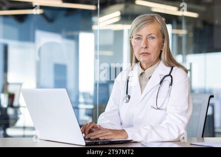 Ritratto di una donna anziana dai capelli grigi seduta in ufficio, che lavora online su un notebook e guarda seriamente nella fotocamera. Foto Stock