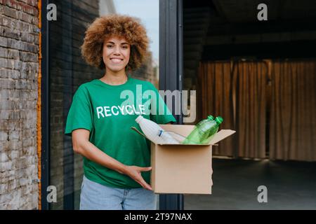 Attivista sorridente che tiene una scatola di cartone con bottiglie di plastica davanti al vetro Foto Stock