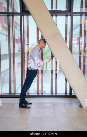 Uomo d'affari che si appoggia alla fronte sulla colonna e utilizza lo smartphone in ufficio Foto Stock