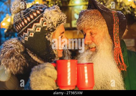 Amici che brindano con VIN brulé o tazze di cioccolata calda al mercatino di Natale, cappello da donna in inverno, uomo con barba bianca Foto Stock