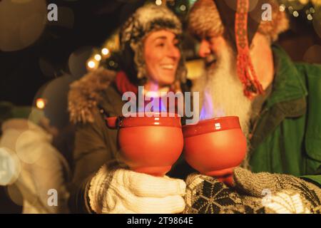 Amici gioiosi che tengono fiammeggianti pinze da fuoco tazze da punch al mercatino di Natale, luci accese e bokeh sullo sfondo. Foto Stock