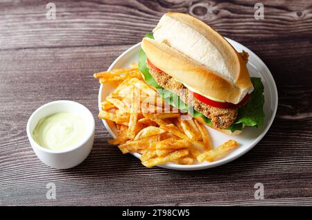 Panino alla milanesa con fette di pomodoro, lattuga, maionese e patatine fritte in un piatto bianco e una ciotola con maionese su fondo di legno. Foto Stock