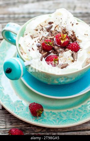 Una bella tazza di caffè blu con panna montata, cacao in polvere e lamponi su sfondo rustico di legno Foto Stock