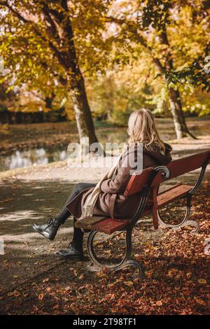 Donna in cappotto che riposa e siede sulla panchina nel parco autunnale. La donna premurosa migliora la sua consapevolezza e la sua salute mentale all'aperto Foto Stock