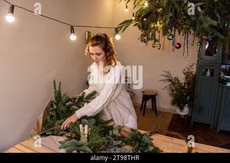 La giovane donna con maglione in maglia leggera sceglie aghi di pino verde naturale per la corona di Natale fatta a mano su un tavolo di legno. La ragazza millenaria fa la decoratio Foto Stock