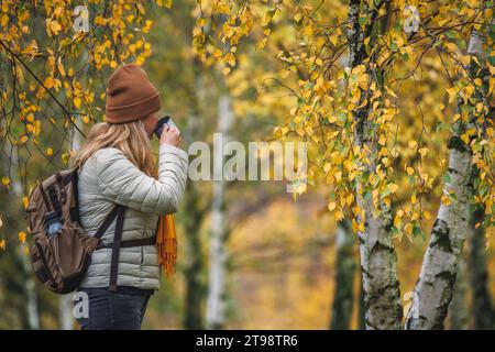 Donna che beve tè caldo dalla tazza da viaggio durante l'escursione nella foresta autunnale. Goditi i boschi nella stagione autunnale Foto Stock