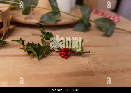 Su un tavolo di legno giace un ciuffo naturale di agrifoglio verde con bacche rosse. Simbolo di Natale. Pianta ornamentale per la realizzazione di bouquet invernali, ghirlande, interni d Foto Stock