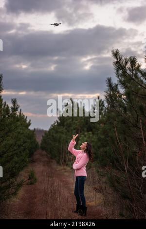 Giovane donna in maglia rosa in campagna tra alberi di conifere nella foresta in posa per una macchina fotografica drone nel cielo. Ragazza riccia che si diverte a nat Foto Stock