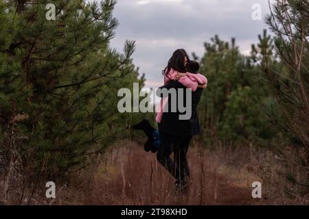 La coppia felice gira nella foresta di conifere lungo il sentiero. Il giovane abbraccia strettamente la donna dai capelli ricci con un maglione rosa. Fidanzamento invernale, matrimonio autunnale. Proposa Foto Stock