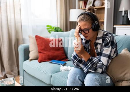 Giovane ragazza triste seduta da sola a casa sul divano ascoltando musica triste con le cuffie wireless che piangono e si puliscono il naso con un fazzoletto di carta. Ricordi un Foto Stock