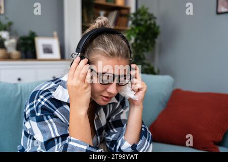 Giovane ragazza triste seduta da sola a casa sul divano ascoltando musica triste con le cuffie wireless che piangono e si puliscono il naso con un fazzoletto di carta. Ricordi un Foto Stock