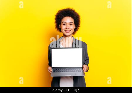Splendida donna afro-americana o brasiliana, studentessa, con i capelli ricci corti, che tiene il laptop, che mostra uno schermo mockup bianco per la pubblicità, stare su sfondo giallo isolato, sorridere Foto Stock