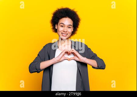 Bella, sincera e piacevole giovane donna afro-americana, in abbigliamento casual, fa gesto cardiaco con le mani, dimostra il segno dell'amore, si trova su sfondo arancione isolato, guarda la macchina fotografica, sorride amichevole Foto Stock