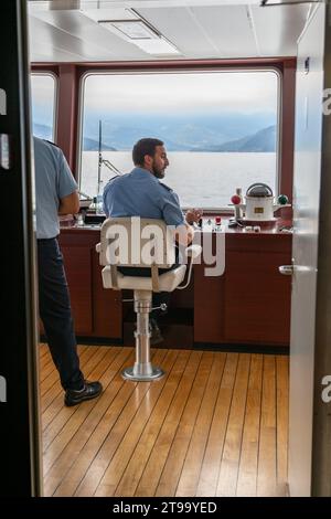 Il capitano di un traghetto navigazione Laghi siede sulla sedia del capitano mentre la barca attraversa il lago di Como, Lombardia, Italia. Foto Stock