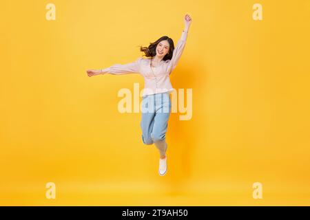 Allegra donna asiatica in abiti casual che sorride e salta con le mani in alto in un colorato giallo sfondo isolato Foto Stock