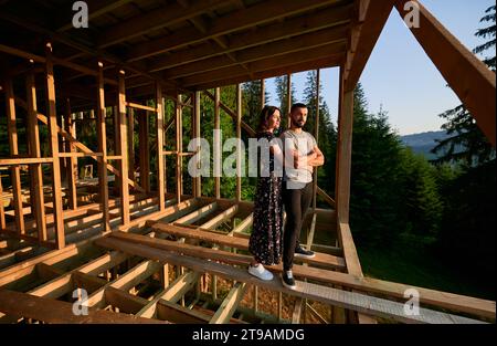 Uomo e donna che ispezionano la futura dimora in legno annidata tra le montagne vicino alla foresta. Coppia giovane in cantiere la mattina presto. Concetto di costruzione ecologica contemporanea. Foto Stock