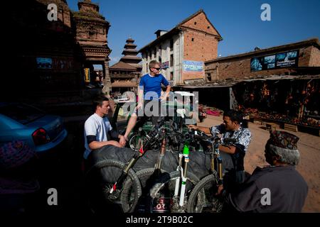 Due uomini viaggiano seduti sul retro di un servizio di prelievo con mountain bike mentre parlano con la gente del posto in una città. Foto Stock