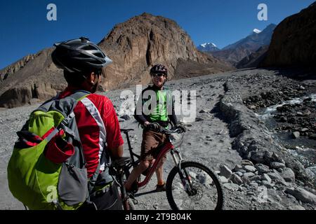 Due uomini conversano in mountain bike vicino al fiume con vista sulle montagne e cielo blu. Foto Stock