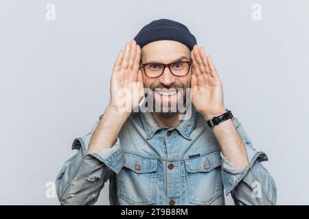 Uomo gioioso con occhiali e berretto, che cova mano a orecchio, in una giacca in denim, su uno sfondo leggero Foto Stock