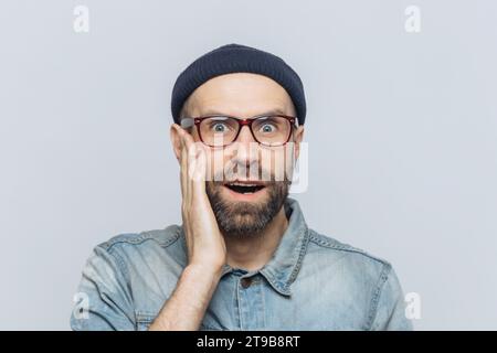 Uomo stupito con una mano sulla guancia, con berretto e occhiali, su uno sfondo grigio chiaro Foto Stock