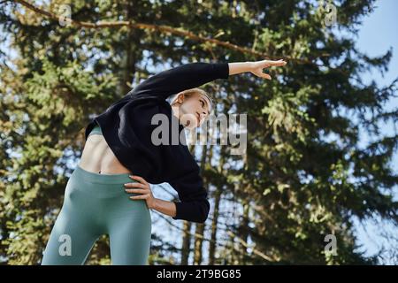 giovane donna sportiva con i capelli biondi e abbigliamento sportivo che allunga il corpo mentre si allena nel parco Foto Stock