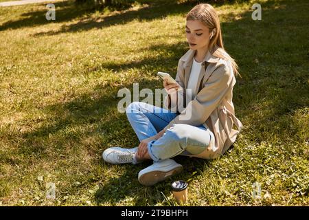 giovane donna bionda con auricolari wireless e trench che utilizza lo smartphone e si siede sul prato Foto Stock