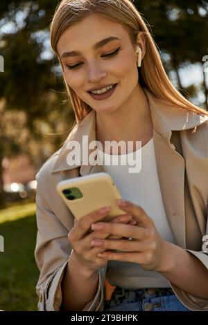 felice donna bionda con auricolari wireless e trench beige che usa il suo smartphone nel parco Foto Stock