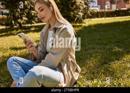 donna bionda felice con auricolari wireless e trench che usa lo smartphone e si siede sul prato Foto Stock