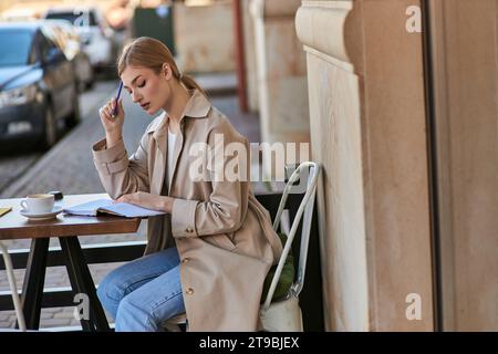 donna pensiva in trench seduta al tavolo con una tazza di caffè e tenendo la penna, scrivendo il diario Foto Stock