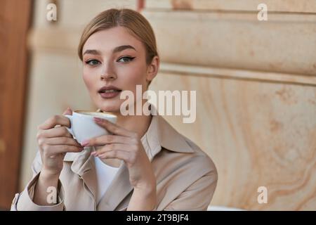 giovane donna elegante con un elegante trench che regge una tazza di caffè mentre si siede in una caffetteria o in una bevanda Foto Stock