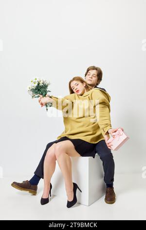 Periodo di bouquet di caramelle. Coppia di abbracci seduti sul palco in una giacca con mazzi di fiori e presenti sullo sfondo bianco dello studio. Foto Stock