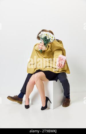 Periodo di bouquet di caramelle. La coppia tiene insieme un mazzo di fiori e un viso coperto mentre si siede in una giacca come un corpo intero contro uno studio bianco Foto Stock