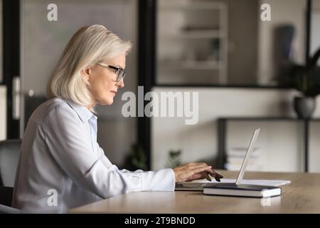 Donna anziana bionda seria che scrive testo sul notebook Foto Stock