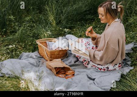 Donna sorridente seduta su una coperta e che mangia panino alla cannella mentre legge un libro Foto Stock