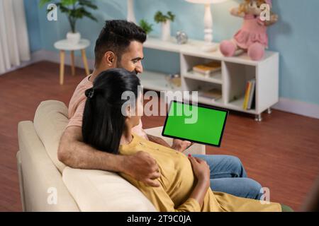 Spalla di una donna incinta indiana con marito che guarda un tablet digitale con schermo verde a casa - concetto di utilizzo di app o applicazione, pubblicità Foto Stock