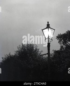 Anni '1950, storico, serale e antico lampione vittoriano o lanterna con lampione in ghisa, illuminato, Inghilterra, Regno Unito. Nel 1807, Pall Mall a Londra, in Inghilterra, vide la prima illuminazione pubblica delle strade, che era generata dal carburante a gas prodotto dal carbone e questa rimase la fonte primaria per le lanterne di strada fino agli anni '1930 Qui sul montante della lampada, una barra orizzontale, che forniva un appoggio sicuro per la scala degli accendini. Foto Stock