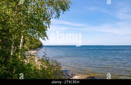 Splendido panorama paesaggistico sul lago e sul lungolago con alberi verdi e spiaggia rocciosa sulla riva, acqua e cielo blu, con poche nuvole sottili. Foto Stock