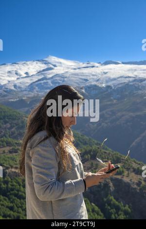 donna latina, dai capelli lunghi con occhiali da sole che guardano lo smartphone, in montagna sullo sfondo della sierra nevada, Foto Stock