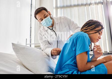 giovane medico indiano che esamina il respiro di malato paziente afroamericano con stetoscopio, assistenza sanitaria Foto Stock