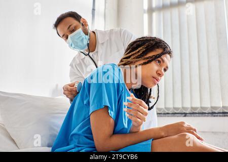 giovane medico indiano che esamina il respiro di malato paziente afroamericano con stetoscopio, assistenza sanitaria Foto Stock