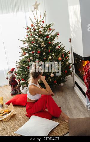 Giovane donna siede accanto a un caminetto luminoso in un accogliente soggiorno adornato con un albero di Natale e decorazioni festive. Foto Stock